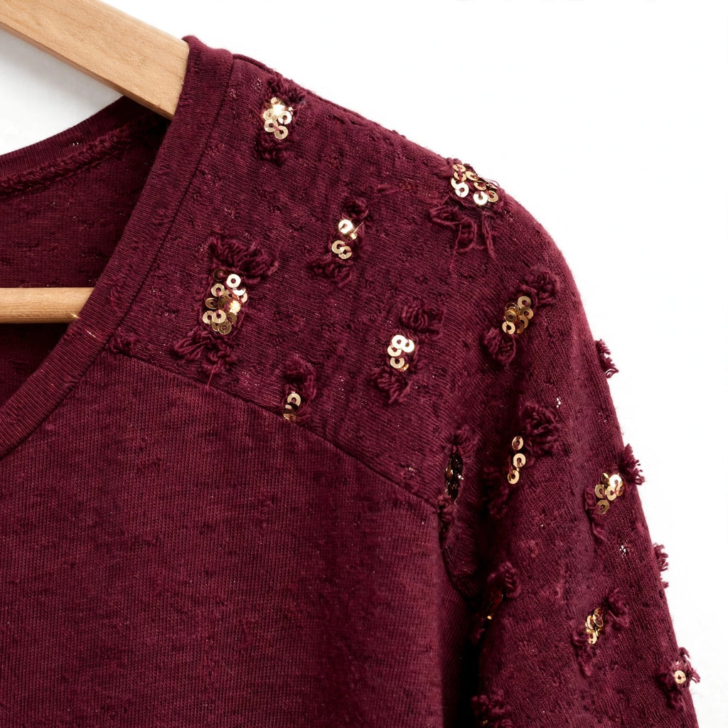 Blusa las cucadas de erisa color burdeos con detalles de lentejuelas doradas en los hombros sobre percha de madera